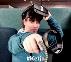 Kuvassa Katja Huttunen VR-lasit ohjallaan katsoo kameraan ja osoittaa ohjaimella sitä kohti.