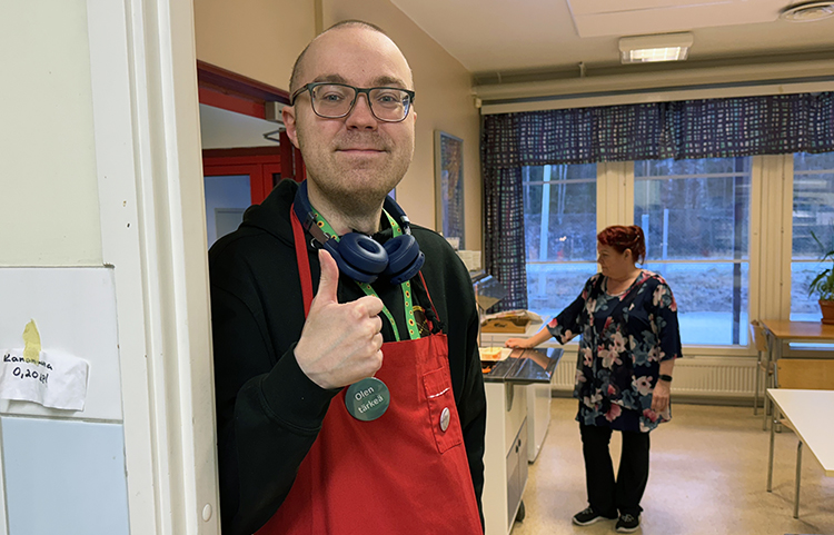 Kaksi henkilöä, etualalla mies punaisessa esiliinassa, peukalo pystyssä.