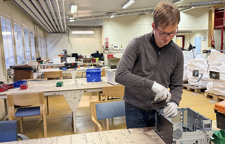Mies työskentelee hanskat kädessä puretun tietokoneen parissa. Taustalla näkyy työpöytiä. 