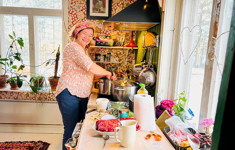 Hymyilevä nainen ison kattilan ääressä viihtyisässä ja värikkäässä keittiössä, jossa on viherkasveja ikkunalaudalla, kukkatapetti taustalla, edessä pöydällä ruokatarvikkeita.