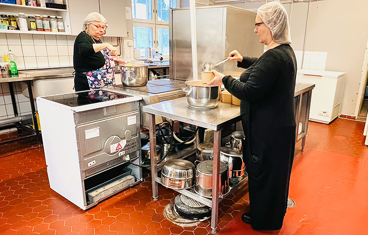 Kaksi naista ravintolakeittiössä, suojapäähineet päässä. He annostelevat ruokaa pahvikippoihin. Pöydän alla näkyy paljon isoja kattiloita.