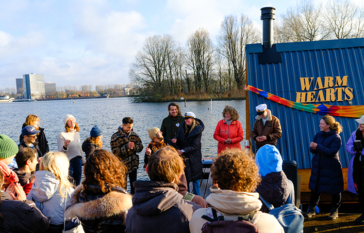 Ihmisjoukko järven rannalla. He seisovat piirissä, osa selin kameraan, osan kasvot näkyvät. Vieressä rakennus, josta näkyy teksti Warm Hearts. Aurinko paistaa, puut ovat lehdettömiä.