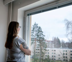 Nainen katsoo ulos ikkunasta, taustalla näkyy kerrostaloja.