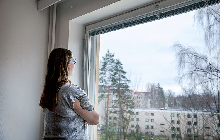 Nainen katsoo ulos ikkunasta, taustalla näkyy kerrostaloja.