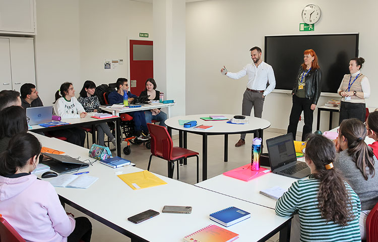 Luokkahuone, jossa oppilaat istuvat ringissä ympäri huonetta, papereita ja vihkoja pöydillä. Luokan edessä kolme henkilöä seisoo.